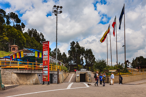 Zipaquira, Colombia - June 16, 2017: Some tourists on the Plaza del Minero near the entrance to the Catedral de Sal, a subterranean church located in an old halite mine, in Zipaquira, in the Cundinamarca Department of the Latin American Country of Colombia. The elevation is about 9000 feet above mean sea level. On the upper level is an area for souvenir shops; flags of the Country, Department and City are seen to the right. A tall signboard advertises the the restaurants just off the Plaza. Photo shot in the morning sunlight; horizontal format. Copy space.