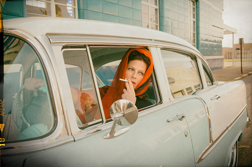 Woman in a red head scarf in her 20s smokes a cigarette and looks out an open car window while driving a vintage pale blue and white car stopped at an intersection, Evansville, IN, USA