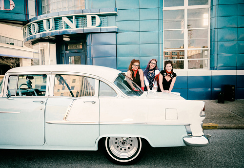Vintage scene of three smiling young women leaning over the back of a restored pale baby blue Bel Aire 1955 Chevy parked on the street at the Greyhound station in downtown Evansville, IN, USA