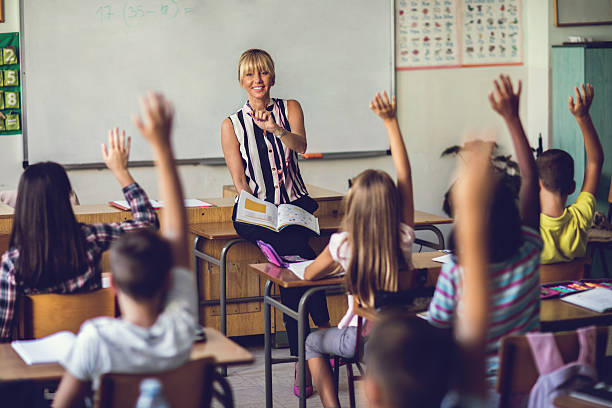 Smiling elementary school teacher asking the question in the classroom. Happy female teacher in the classroom pointing at school kids who are raising hands to answer the question. elementary student pointing stock pictures, royalty-free photos & images
