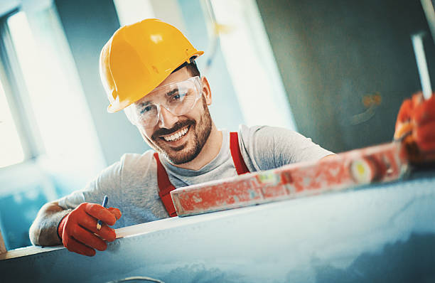 Cheerful construction worker. Closeup front view of a cheerful handsome construction worker leaning against a white wall and looking at camera. He's wearing red protective gloves and yellow work helmet. repairperson occupation stock pictures, royalty-free photos & images