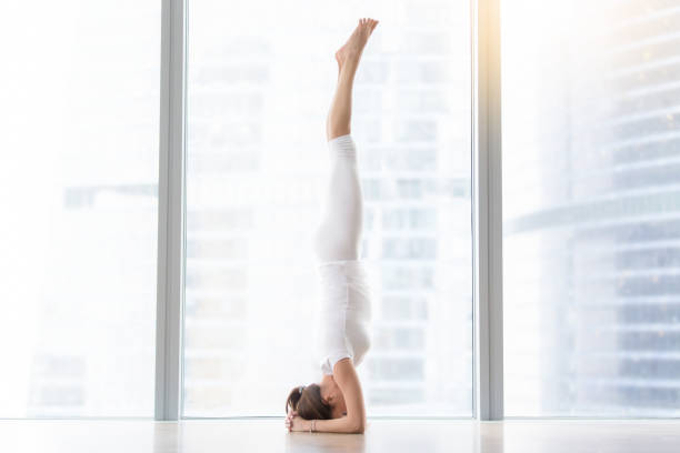 Young attractive woman in salamba sirsasana pose near floor wind Young attractive woman practicing yoga, standing in salamba sirsasana exercise, headstand pose, working out, wearing sportswear, white t-shirt, pants, full length, near floor window, horizontal shirshasana stock pictures, royalty-free photos & images