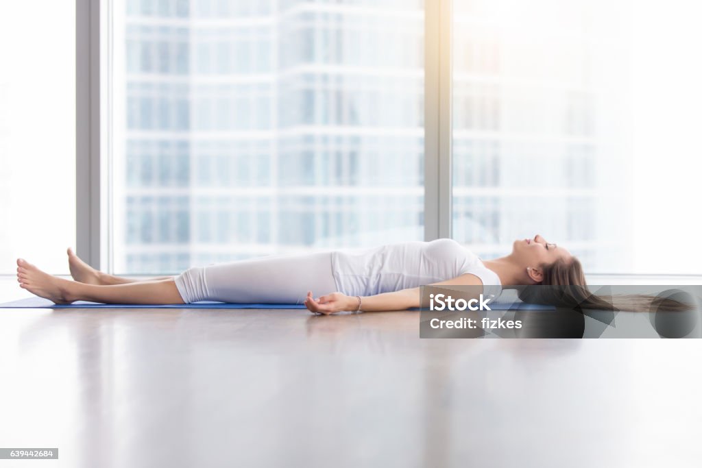 Young attractive woman in Corpse pose against floor window Young woman practicing yoga, lying in Savasana exercise, Dead Body, Corpse pose, working out, wearing sportswear, white t-shirt, pants, indoor full length, near floor window with city view, side view Yoga Stock Photo