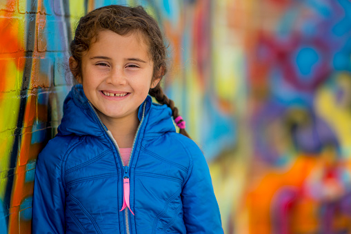 An elementary age girl is standing outside in an urban city - a colorful graffiti wall is in the background. She is smiling and looking at the camera.