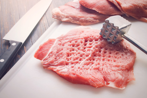 Tenderized piece of raw meat on a white cutting board, with knife and tenderizer