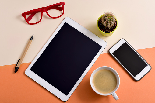 high-angle shot of an office desk full of things, such as a pair of red eyeglasses, a cup with white coffee, a tablet, a smartphone, a pen and a cactus
