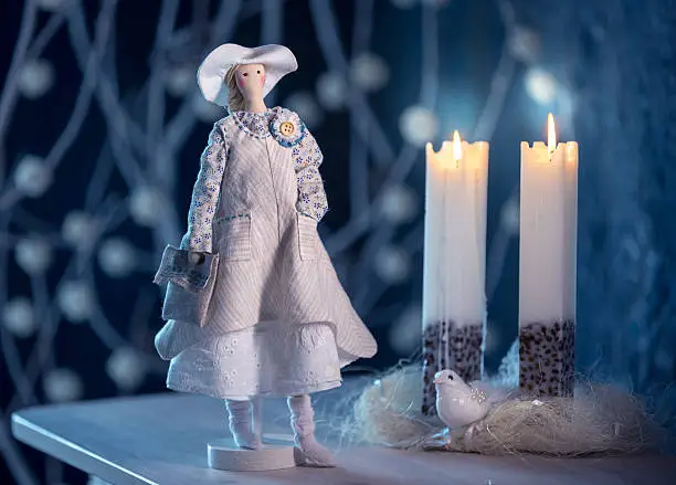 Handmade rag-doll. Studio shooting. Doll girl in hat and in white sundress. Textile tilda doll standing on the shelf next to the candlestick with two burning candles