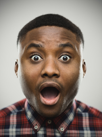 Close-up portrait of shocked man. Amazed male is raising eyebrows against gray background. He is wearing plaid shirt. Vertical studio photography from a DSLR camera. Sharp focus on eyes.