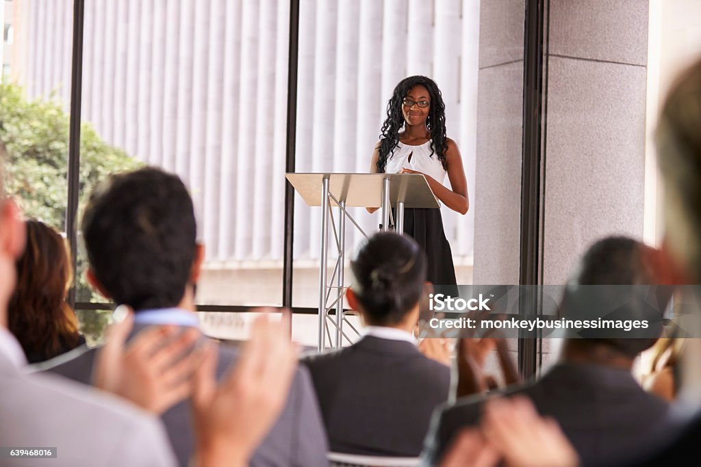 Pubblico al seminario che applaude giovane donna di colore al leggio - Foto stock royalty-free di Oratore pubblico