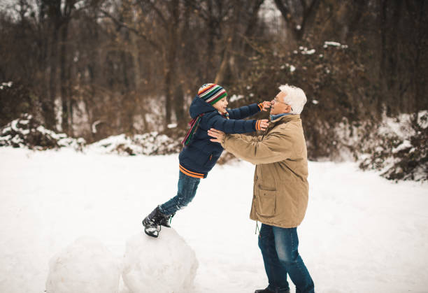 A photo of a grandfather and grandson playing in the snow. They have winter clothing. The kid is jumping and grandfather is helping him to land.