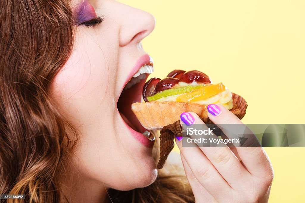 Closeup woman eating fruit cake sweet food Sweetness and happiness concept. Closeup cute woman curly hair eating fruit cake cupcake face profile yellow background Adult Stock Photo