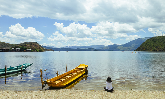 Single young Chinese woman looks into skyline, sitting on bank of Lugu lake, Lijiang, China, back to camera
