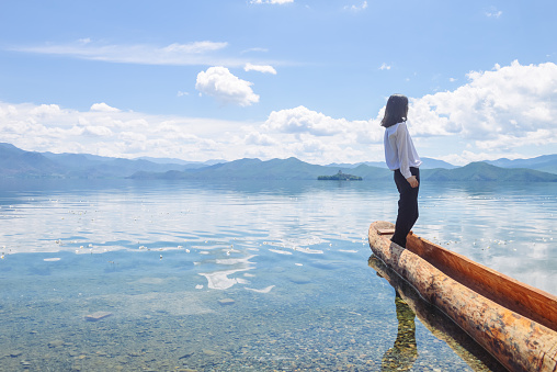 Single young Chinese woman looks aside, back to camera, while standing on boat by the bank of Lugu lake, Lijiang, China