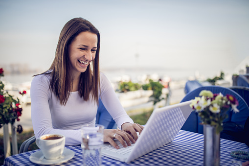 Young brunette woman enjoying the sunny morning at the beach restaurant with a cup of coffee and a laptop computer.