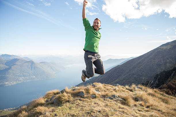 Young hiking man at the top of a mountain jumping high up for success. Swiss Alps and lake on the background. Autumn season.
