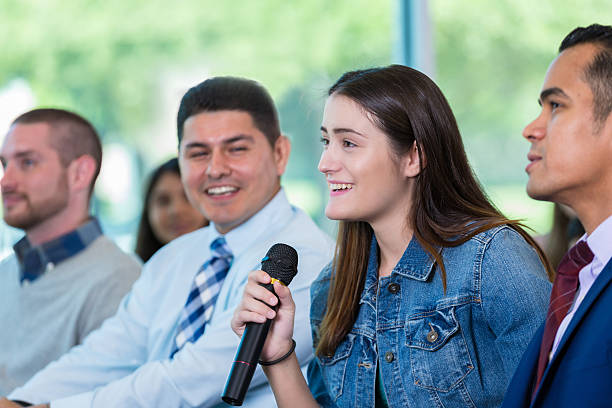 Young woman asks question during town hall meeting Confident young Caucasian woman asks question during town hall meeting. She is holding a microphone. constituency photos stock pictures, royalty-free photos & images