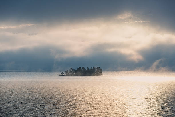 Clouds and fog lifting to reveal a tiny island in the sunlight on Flathead Lake in Montana, USA. No people in this high resolution color photograph with plenty of copy space and a horizontal composition. Beautiful nature background.