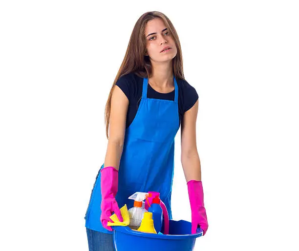 Tired young woman in blue T-shirt and apron with pink gloves holding cleaning things in blue washbowl on white background in studio
