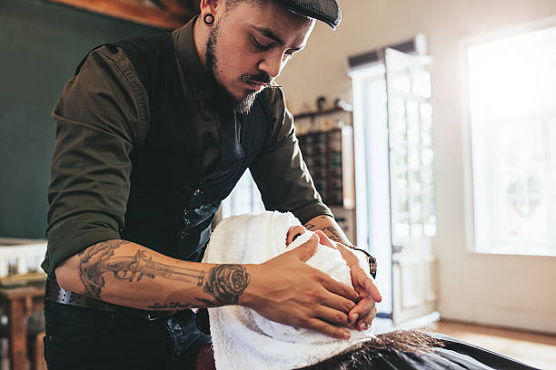 Hipster barber applies a warm cloth on face of male client. Hairdresser covering face of male client with towel.
