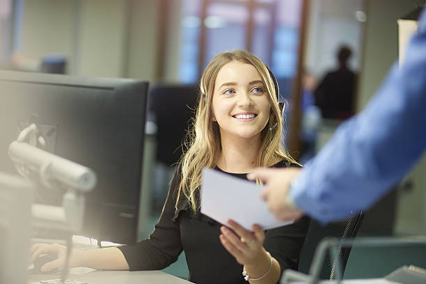 enjoying the new job a young woman sits at her desk and is handed some paperwork by a colleague out of shot. She is smiling and enjoying her placement at the large open plan office . She is wearing a headset . first job photos stock pictures, royalty-free photos & images