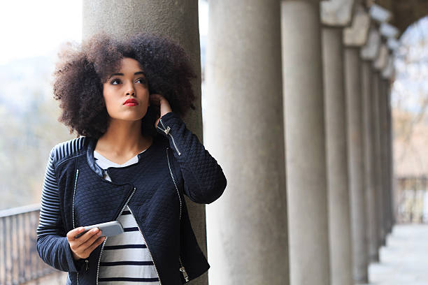 Beautiful young woman using smart phone on street. Looking up, one hand on head. Wears casual clothes. Ancient columns in row on background.