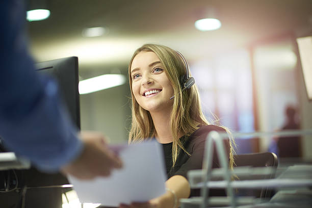 happy office intern a young woman sits at her desk and is handed some paperwork by a colleague out of shot. She is smiling and enjoying her placement at the large open plan office . She is wearing a headset . first job photos stock pictures, royalty-free photos & images