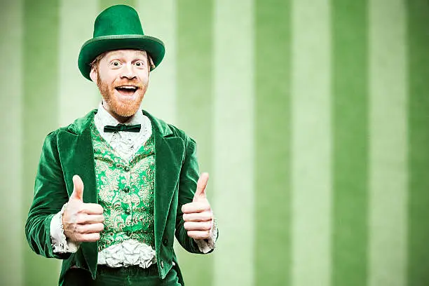 A stereotypical Irish character all ready for Saint Patricks day.  He gives two thumbs up, a big smile on his face.  Studio portrait.  Horizontal image with copy space.  Green striped wallpaper background.