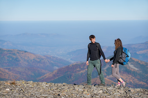 Young couple of tourists climbing up with backpacks on the ridge of the mountain and looking to each other, open overview on the mountains on the background. Couple is holding hands. Side view