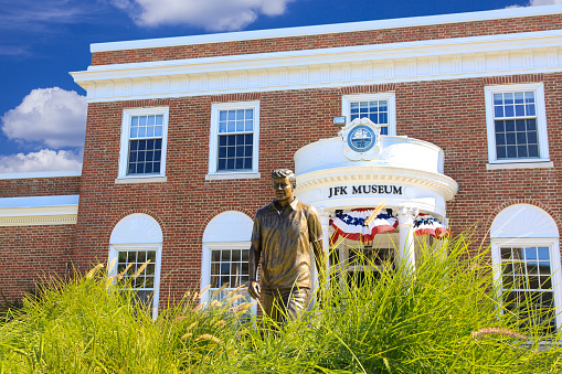 Hyannis, USA - September 6, 2009: John Fitzgerald Kennedy (JFK) Museum in Hyannis, Massachusetts, USA. In foreground is a bronze statue of the JFK walking through green grass. Red brick building with white portico and vivid blue sky with clouds are in the image.