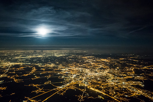 The city Liège in Belgium in the foreground, its lights glowing brightly, as the Moon rises behind thin clouds in the distance
