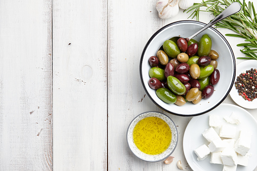 Greek cuisine ingredients: fresh olives mix, feta cheese, rosemary twigs, garlic, peppercorns and olive oil on white wooden background