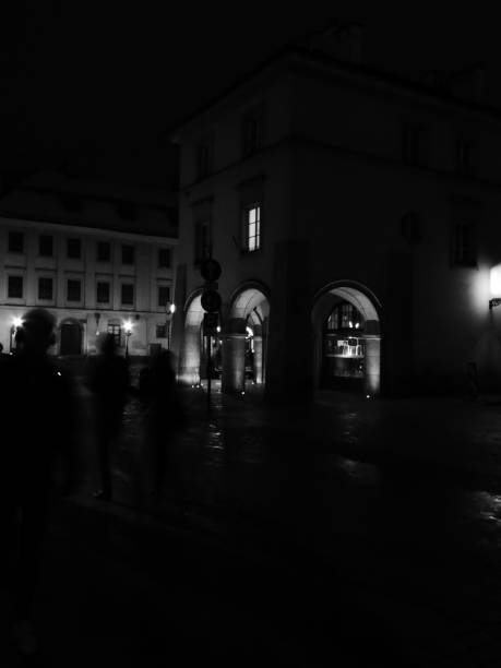 Monochrome shadowy pedestrians in Krakow dimly lit at night. Lights in shop windows.