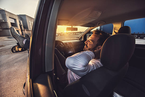Asian large build man wearing black t-shirt and eyeglasses falling asleep while waiting for friend in a car so he's grabbing a pillow and napping with arms crossed on driver's seat.
