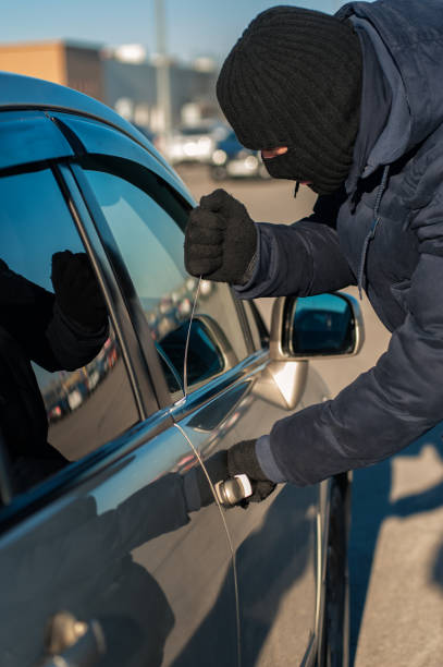 Male thief in black robbery mask breaking into car with screwdriver