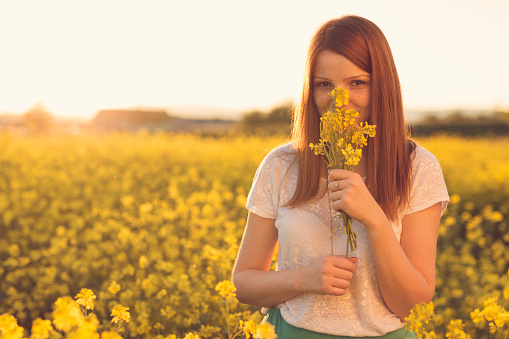 Beautiful, russet young woman standing in the middle of the flower meadow, holding and smelling a bouquet of yellow flowers