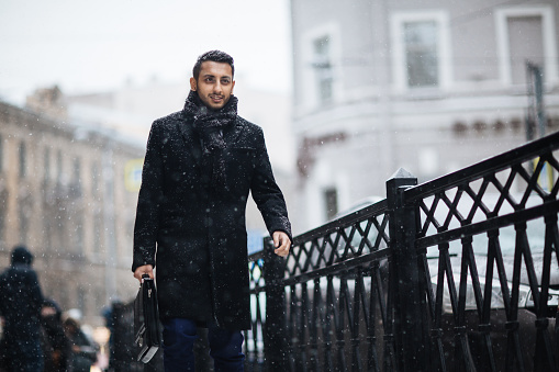 Portrait of modern young Middle Eastern businessman in the streets of snowy big city, he is passing bridge enjoying walk and smiling