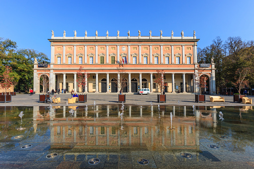 Reggio Emilia, Italy - October 30, 2014: People strolling next to the Teatro Municipale Romolo Valli (Municipal Theatre), built in the mid-1800s. Today the theatre hosts a opera and concert season, as well as a rich programme of ballet. It is located in the city center, surrounded by the public gardens.