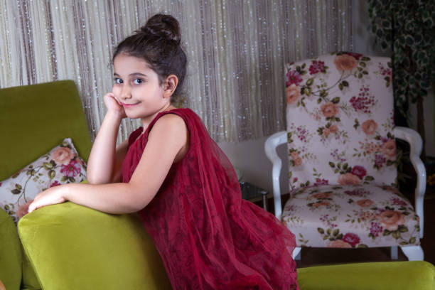 Cute lovlely middle eastern girl with dark red dress and collected hair posing and liying on green sofa at home interior. studio shot.