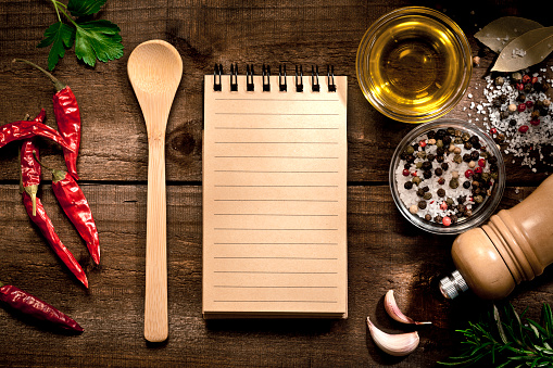 Top view of a blank recipe book with herbs and spices on rustic wooden table. The recipe book is at the center of an horizontal frame with a wooden spoon at the left while the herbs and spices like dried chili peppers, salt, peppercorns, garlic, bay leaves, parsley and rosemary are around it. Copy space available on the recipe book. Predominant color is brown. DSRL studio photo taken with Canon EOS 5D Mk II and Canon EF 100mm f/2.8L Macro IS USM
