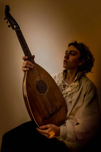 Young man with long blond hair sitting in dark environment and hold in His Hands an Old Oud, Guitar Lute and looking at it. He is dressed in white rustic baroque shirt with ruffle edging to collars.