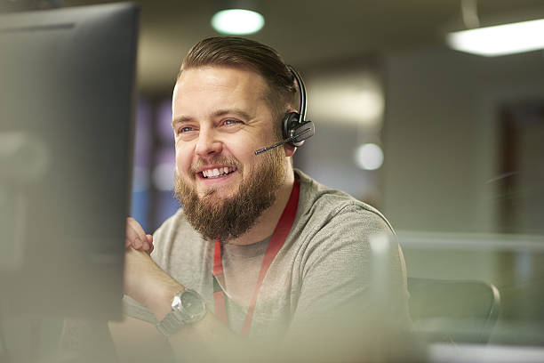 helpful customer service a call centre phone operative in his mid 30s chats on the phone at his desk . He is explaining something to the person on the phone in a friendly manner . behind him a defocussed office interior can be seen . This could be a call centre or an office worker chatting to a customer . customer service stock pictures, royalty-free photos & images