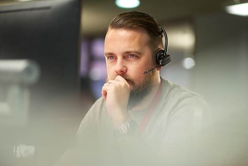 a call centre phone operative in his mid 30s chats on the phone at his desk . He is explaining something to the person on the phone in a friendly manner . behind him a defocussed office interior can be seen . This could be a call centre or an office worker chatting to a customer .