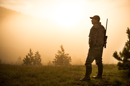 Portrait of a hunter with sniper rifle on the mountain hill. Bright sunlight is breaching through the fog behind him.