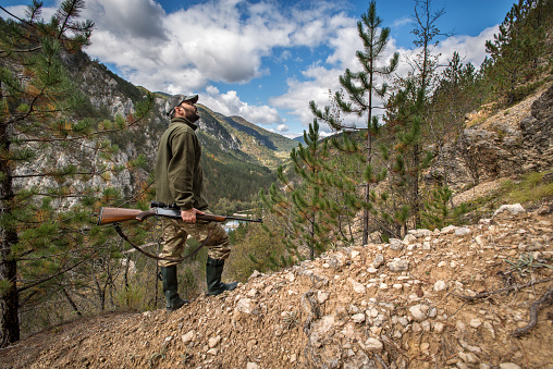 Hunter with sniper rifle standing on steep hill and looking for animal tracks.