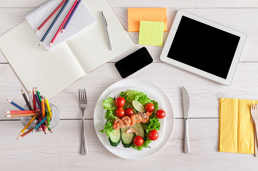 Healthy business lunch in the office, top view of dish on white wooden desk near tablet, mobile phone and open organizer. Salad plate with salmon. Snack at break time