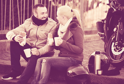Young smiling adults drinking coffee and chatting near motorcycle