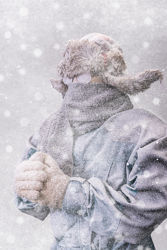 A frozen man with his hands clasped all bundled up in a fur trappers hat, scarf, parka and knit gloves, covered in snow and frost trying to stay warm on a very cold Winter's day as a blizzard's  snow falls around him.
