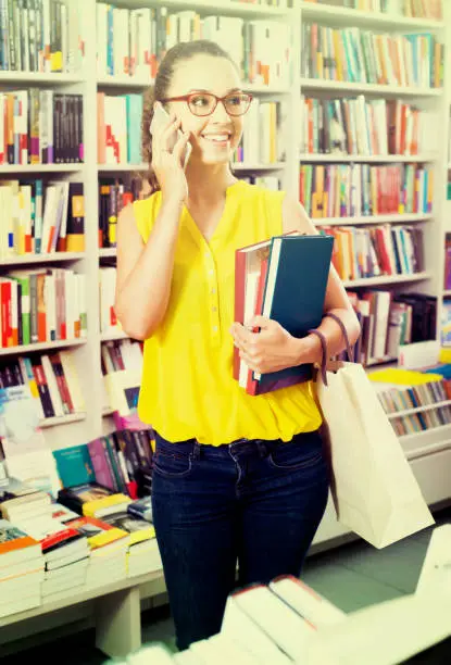 portrait of joyful young woman in glasses picking textbooks for school while talking on telephone in shop