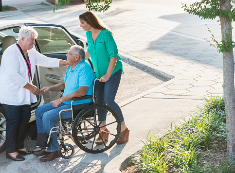 A senior man smiling as he goes from the back seat of car into a wheelchair. He is sitting in the wheelchair, smiling at his wife standing in front of him. His adult daughter is behind the wheelchair.