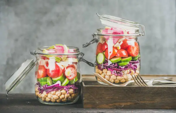 Healthy take-away lunch jars. Vegetable and chickpea sprout vegan salad in glass jars, grey concrete wall background, copy space, selective focus. Clean eating, vegetarian, raw, detox, dieting concept
